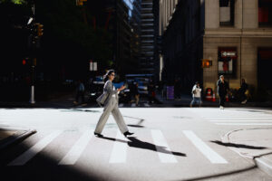 Young businesswoman walking confidently across a bustling New York City crosswalk, focused on checking her smartphone amidst the urban scene